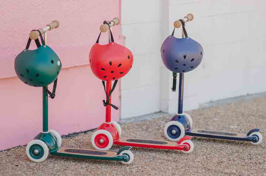 banwood scooters in green, red and blue lined up with matching kids bike hedlmets hanging from handlebars