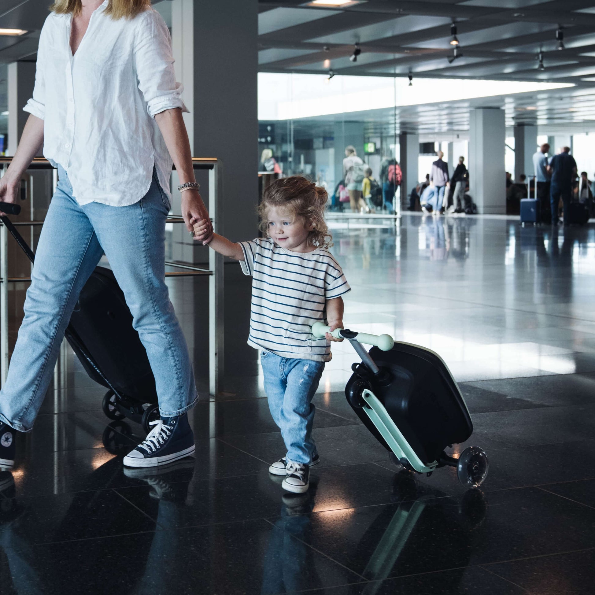 woman holding hands with child who is wheeling Micro Scooter Mini Mint Luggage Scooter through airport