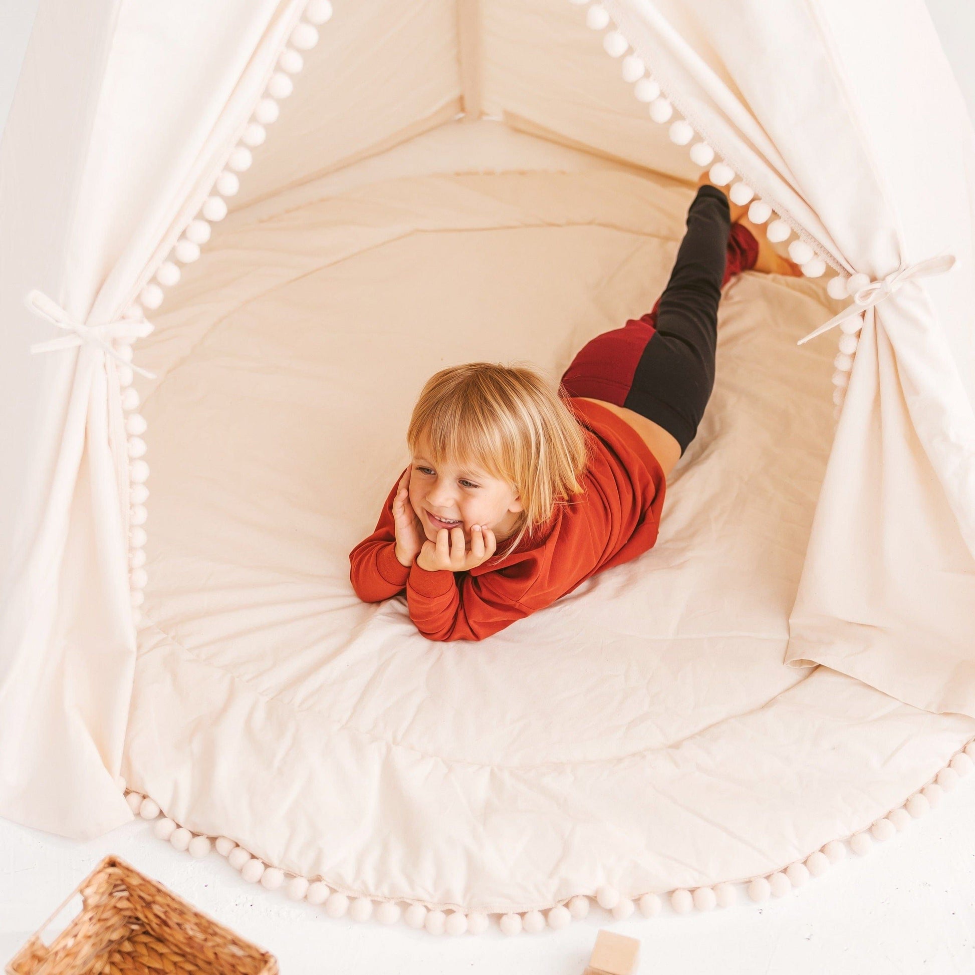 smiling child lying in MINICAMP Extra Large Kids Teepee Tent With Pom Pom Decor