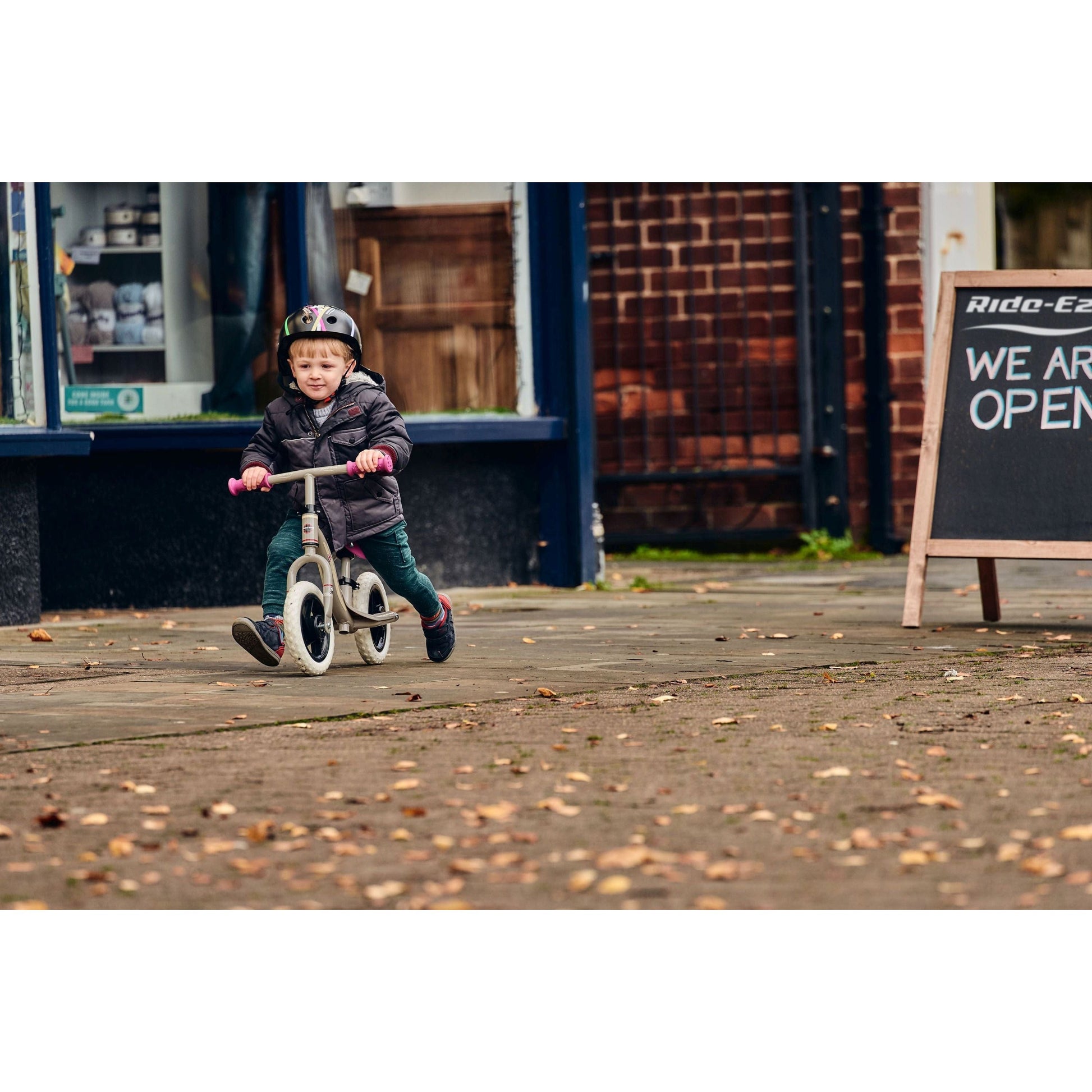 boy riding Ride-Ezy Go Balance Bike - Pink & Silver on pavement in front of shop