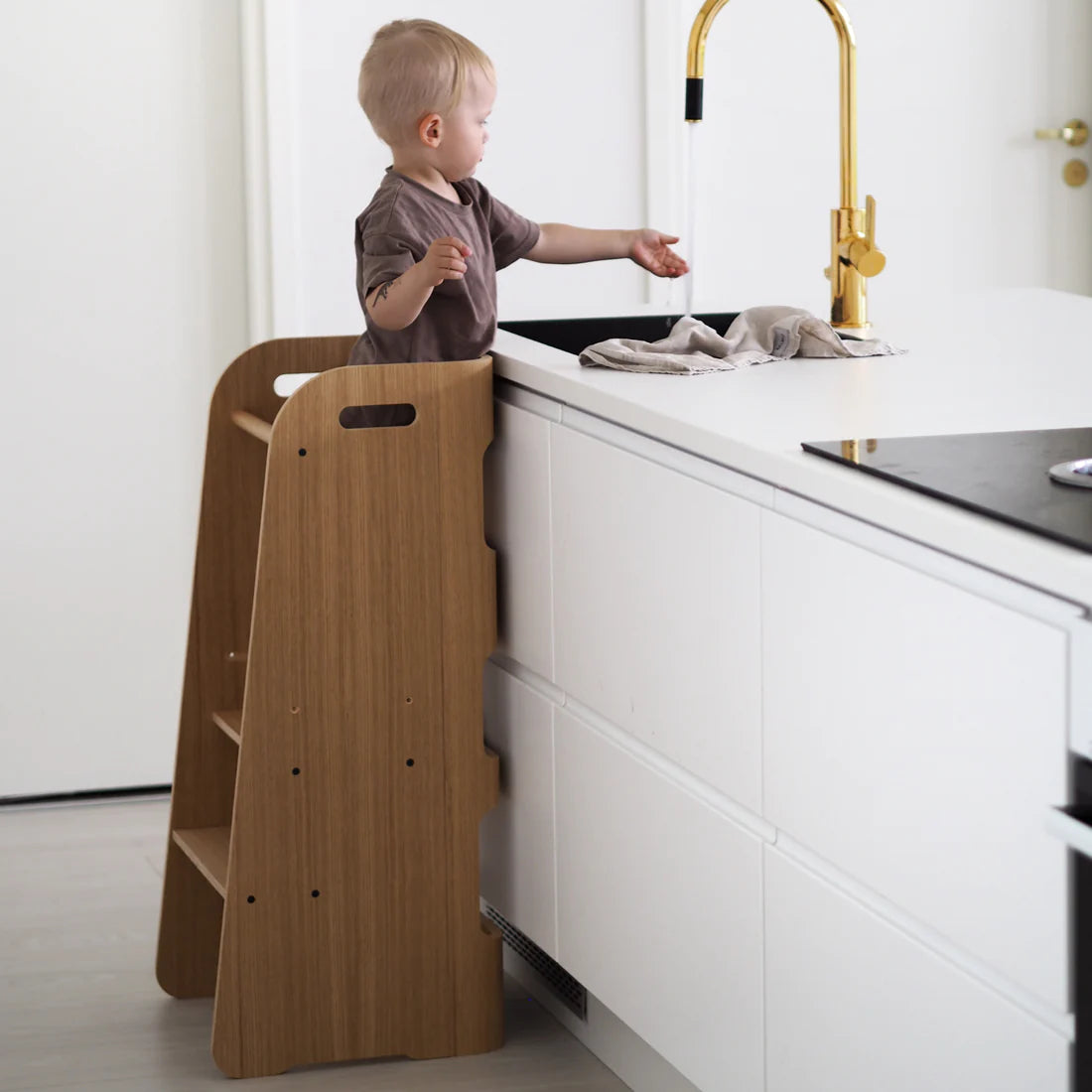 little boy standing on curve lab kitchen helper learning tower and running his hand under tap