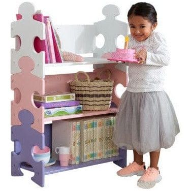 girl holding ckae in front of Kidkraft Puzzle Bookshelf - Pastel