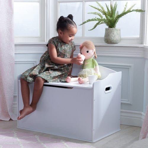 girl sitting on KidKraft Austin Toy Box - White while feeding doll