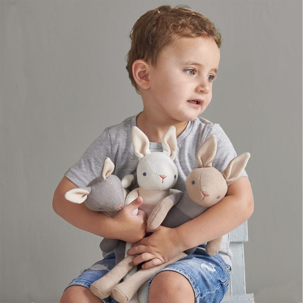 boy sitting on chair holding ThreadBear Baby Comforter, Rattle & Doll Bundle in Cream