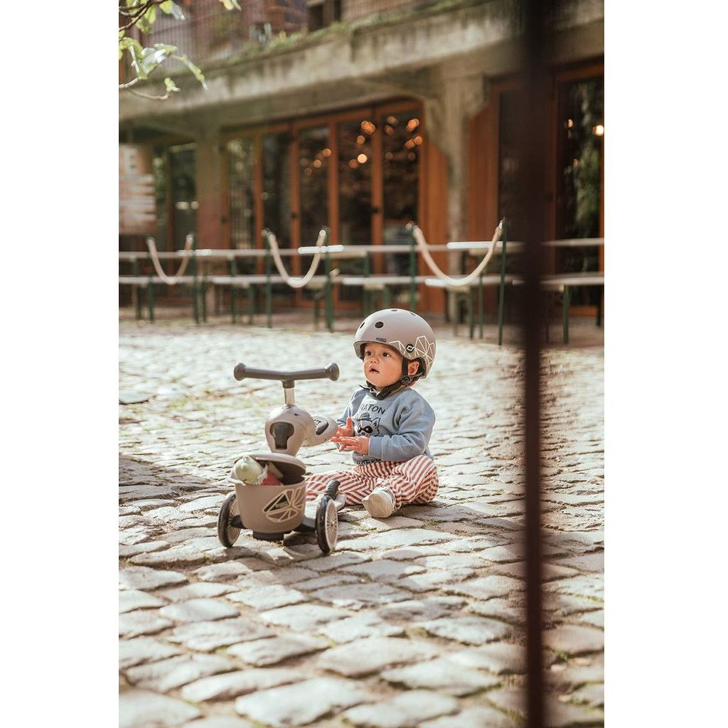 child sitting on cobbled street next to Scoot and Ride Highwaykick 1 Lifestyle Scooter- Brown Lines 