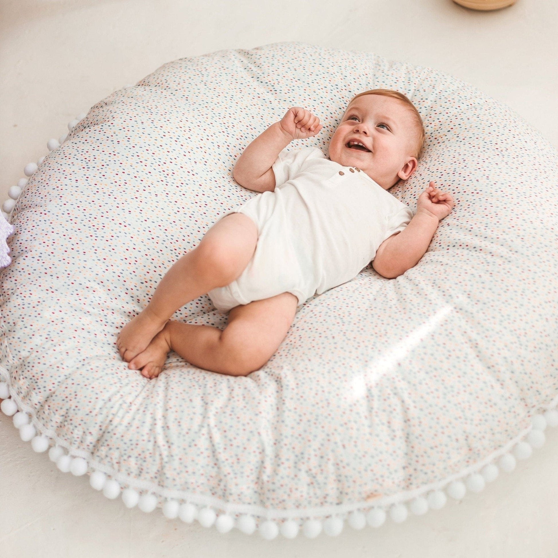 smiling baby lying on MINICAMP Big Floor Cushion With Pompoms in Colour Drops on White