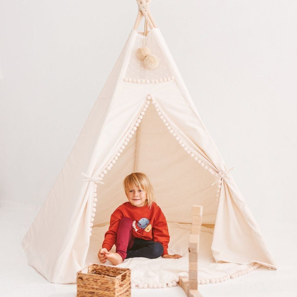 child sitting in MINICAMP Extra Large Kids Teepee Tent With Pom Pom Decor